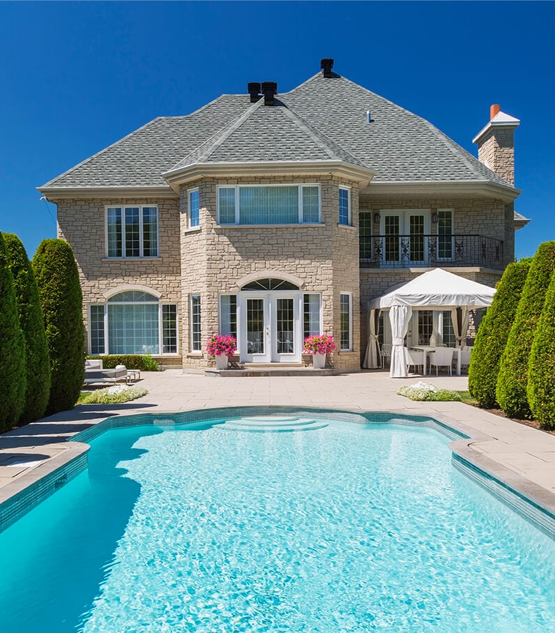 Armonical shot of a pool on a sunny day, surrounded at each side by shrubs of different sizes, and a beautiful gray house
