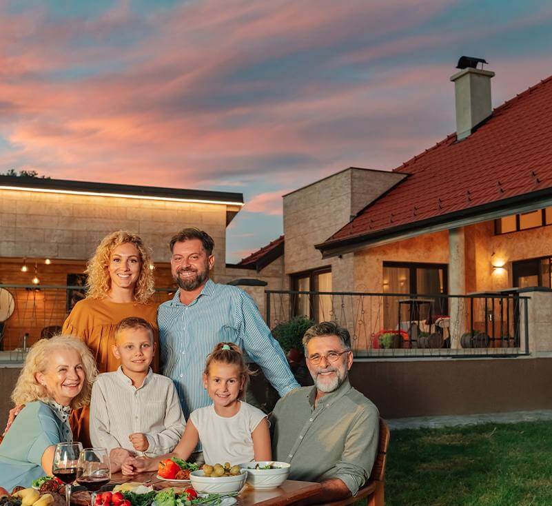 Happy multi-generational family having dinner outside at sunset, roof of home visible in background